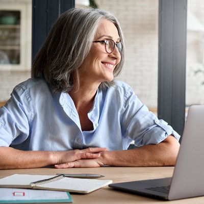 Eine ältere Frau mit Brille sitzt an einem Tisch mit einem Laptop.