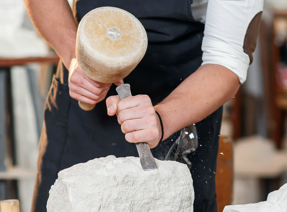 Eine Person schlägt mit einem Holzklotz eine Steinskulptur zurecht. 