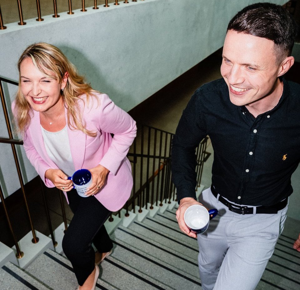 Eine Frau mit rosa Blazer und ein Mann mit schwarzem Hemd gehen lachend die Treppe hoch.