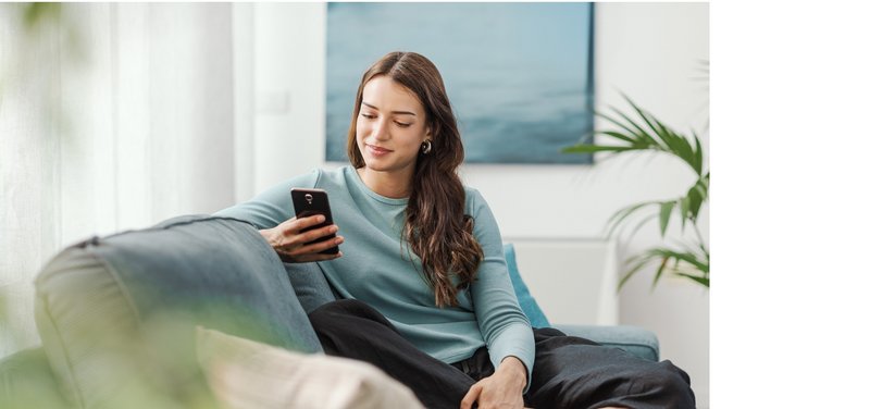 Junge Frau sitzt entspannt auf einem Sofa und schaut auf ihr Handy.