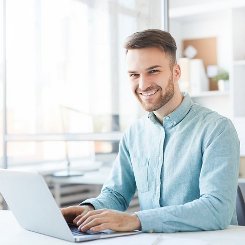 Ein lachender Mann sitzt am Schreibtisch und tippt auf seinen Laptop.