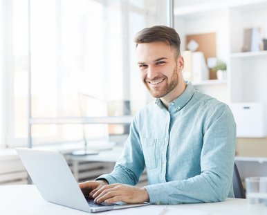 lachender Mann sitzt am Schreibtisch und tippt auf seinen Laptop.