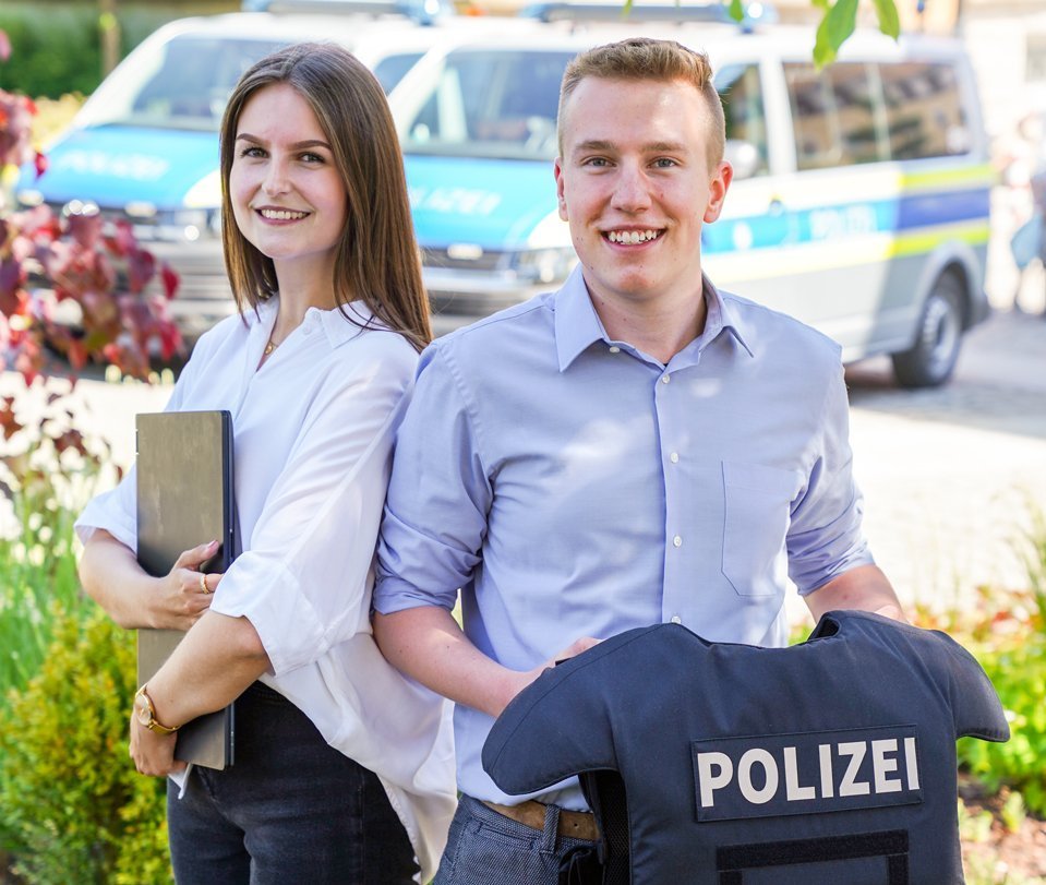 Eine lachende Frau und ein lachender Mann stehen vor einem Polizeiwagen.