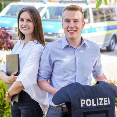 Eine lachende Frau und ein lachender Mann stehen vor einem Polizeiwagen.