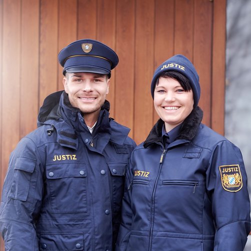 Eine lachende Frau und ein lachender Mann in Justizuniform stehen vor einem Tor.