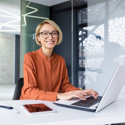 Eine lachende Frau sitzt am Schreibtisch und tippt auf ihren Laptop.