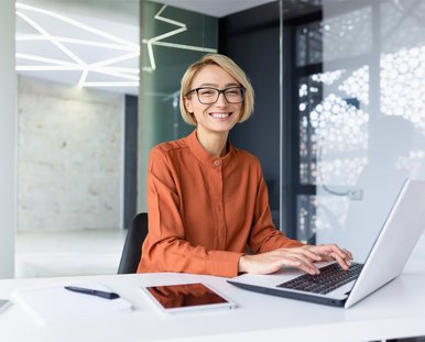 Eine lachende Frau sitzt am Schreibtisch und tippt auf ihren Laptop.