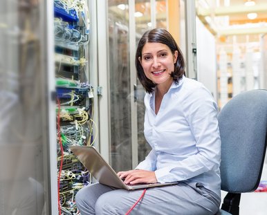 Eine lachende Frau sitzt vor einem Serverraum mit einem Computer.