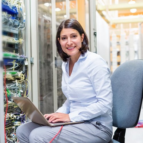 Eine lachende Frau sitzt vor einem Serverraum mit einem Computer.