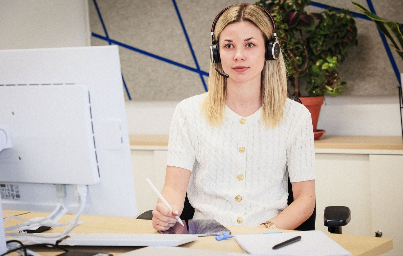 Junge Frau sitzt am Schreibtisch mit Kugelschreiber in der Hand und Headset auf dem Kopf.