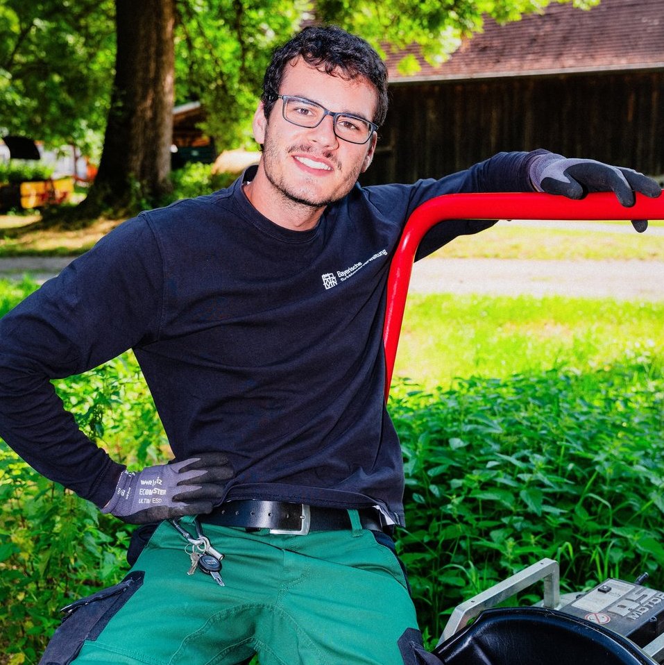 Junger Mann mit Brille lehnt sich entspannt an den roten Griff eines Gartengeräts. Er trägt ein dunkles Langarmshirt, eine grüne Arbeitshose und Handschuhe.
