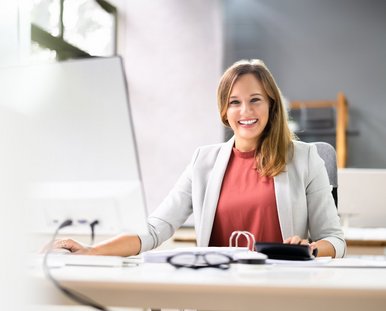 Eine lachende Frau sitzt vor einem Computer an einem Schreibtisch.