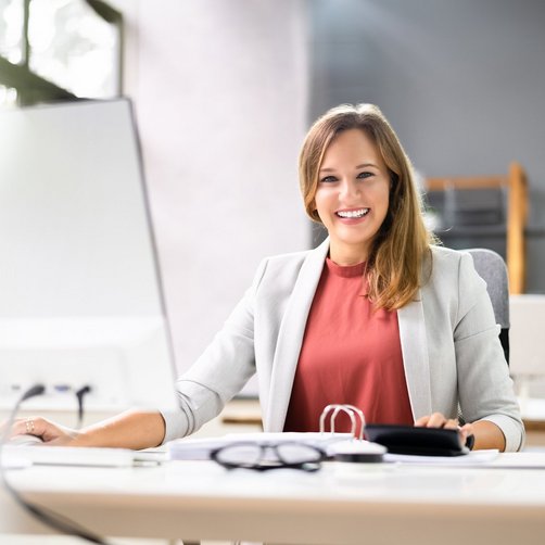 Eine lachende Frau sitzt vor einem Computer an einem Schreibtisch.