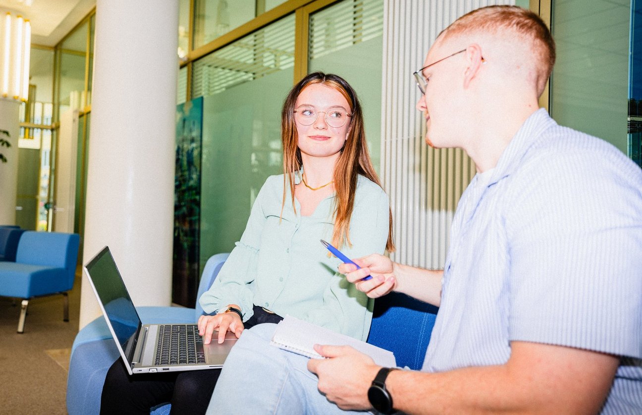 Eine Frau mit Laptop und ein Mann mit Stift sitzen auf einem Sofa und unterhalten sich in einem Bürobereich.