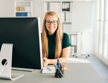 Eine Frau mit Brille sitzt an einem Schreibtisch mit einem Computer.