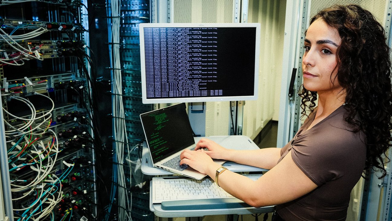 Frau mit dunklen, lockigen Haaren steht im Serverraum vor einem Laptop und Bildschirm.