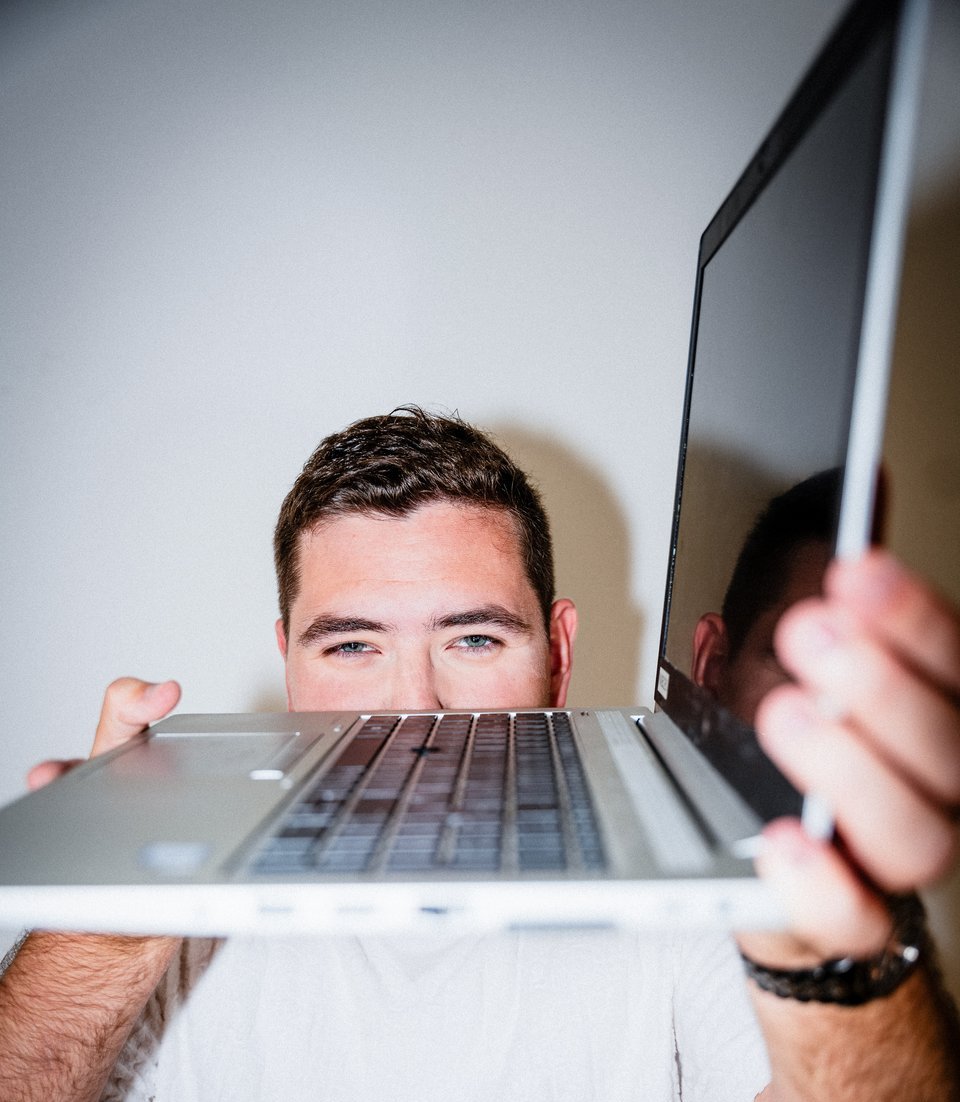 Junger Mann mit weißem T-Shirt und schwarzen, kurzen Haaren hält silbernes Notebook in die Kamera.