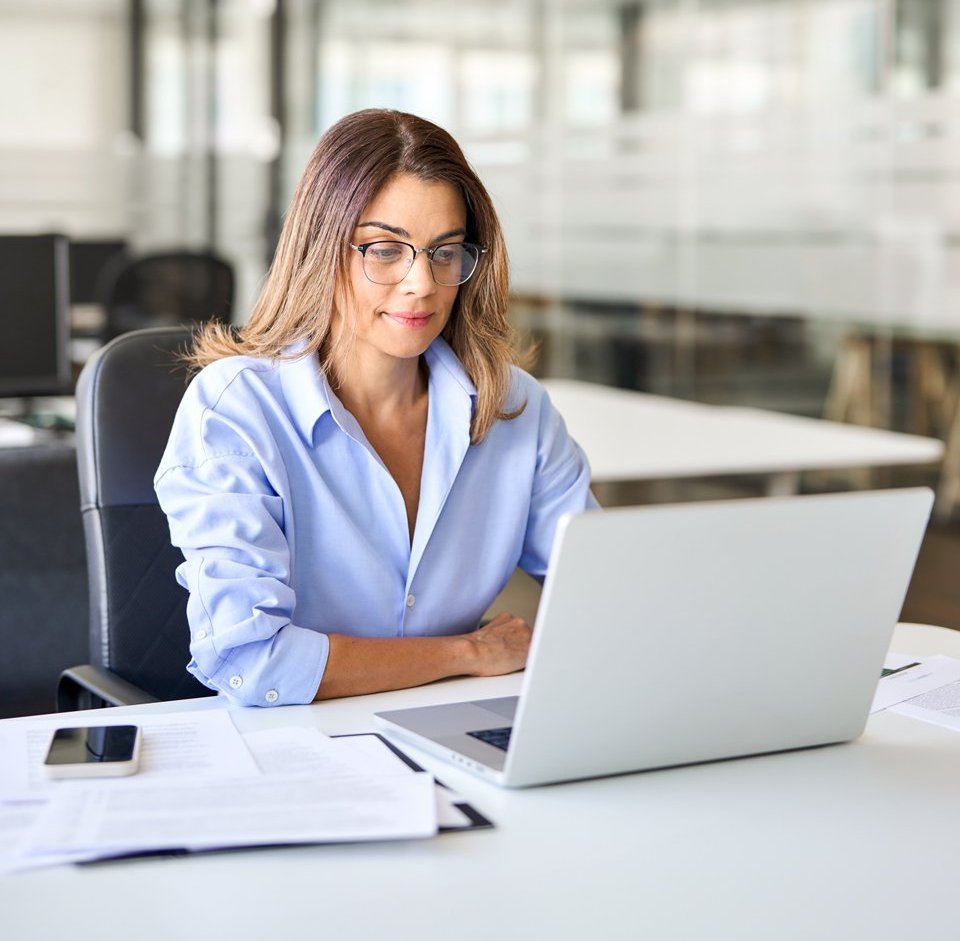 Eine Frau mit Brille sitzt an ihrem Schreibtisch und arbeitet an ihrem Laptop.