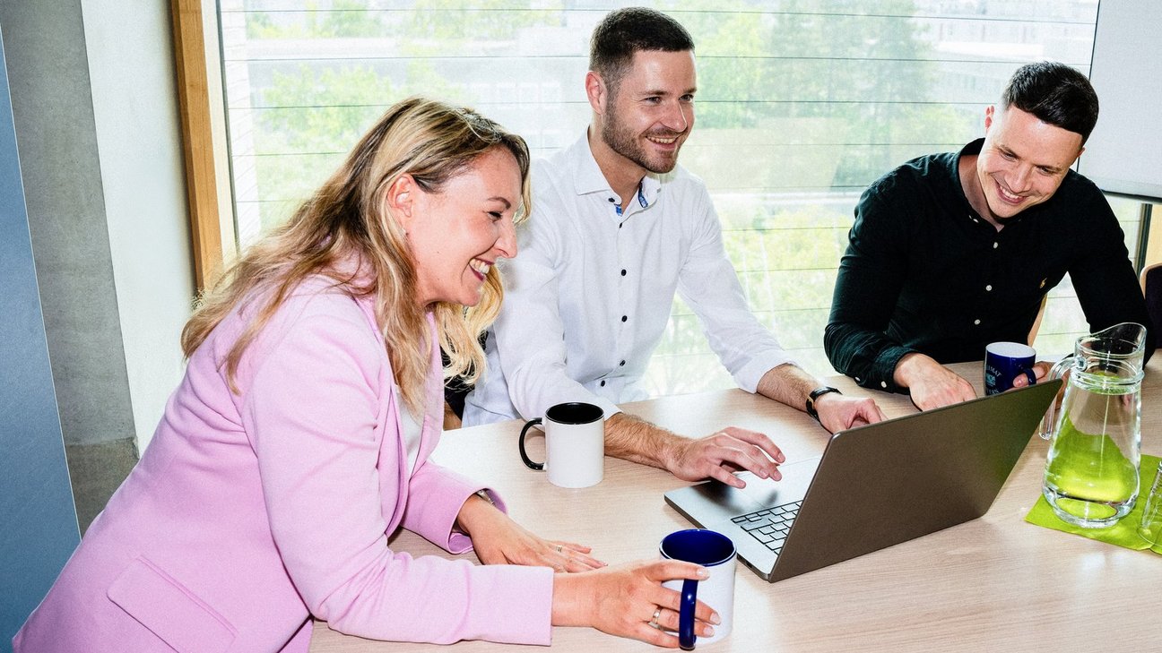 Eine Frau und zwei Männer sitzen an einem Besprechungstisch und schauen gemeinsam in einen Laptop-Bildschirm.