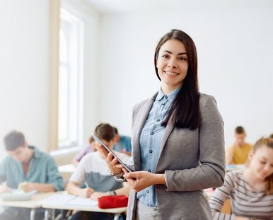 Eine lachende Frau steht vor einer Klasse und hält ein Tablet in der Hand.hält 