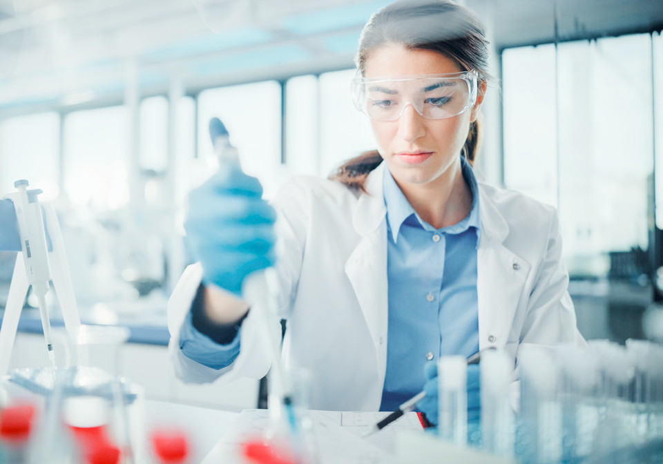 Eine Frau in Laborkittel mit einer Schutzbrille arbeitet mit einem Reagenzglas.