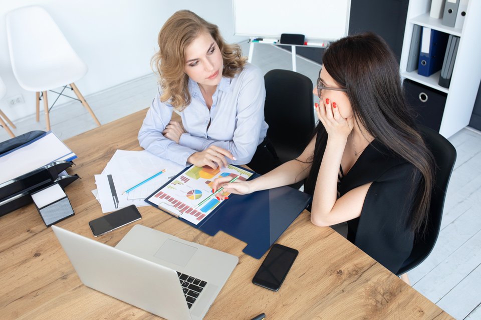 Zwei Frauen sitzen an einem Schreibtisch, darauf ein Computer und Zettel.