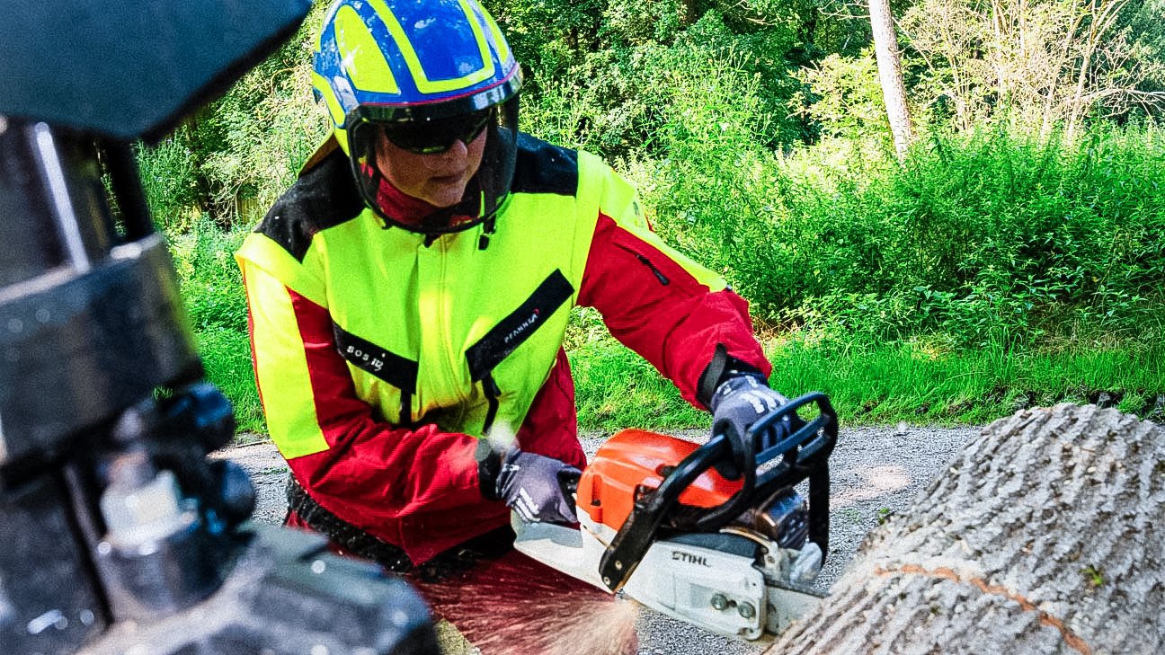 Gärtnerin mit Schutzkleidung schneidet mit Kettensäge Baumstamm.