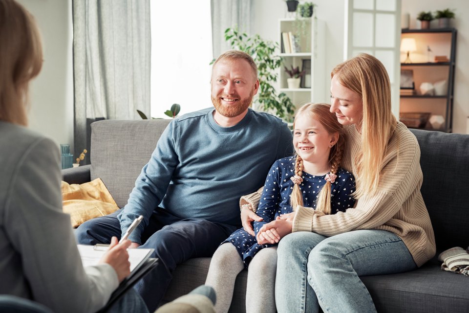 Eine Familie sitzt lachend auf der Couch und unterhält sich mit einer weiteren Person.