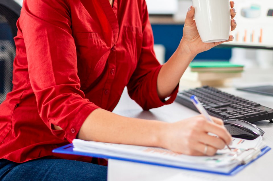 Eine Person schreibt etwas auf einen Zettel und hält eine Kaffeetasse in der anderen Hand.