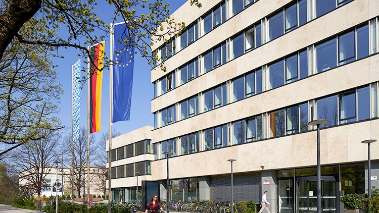 Ein Gebäude großen Fenstern neben einer Deutschland-, Bayern-, und EU-Flagge.