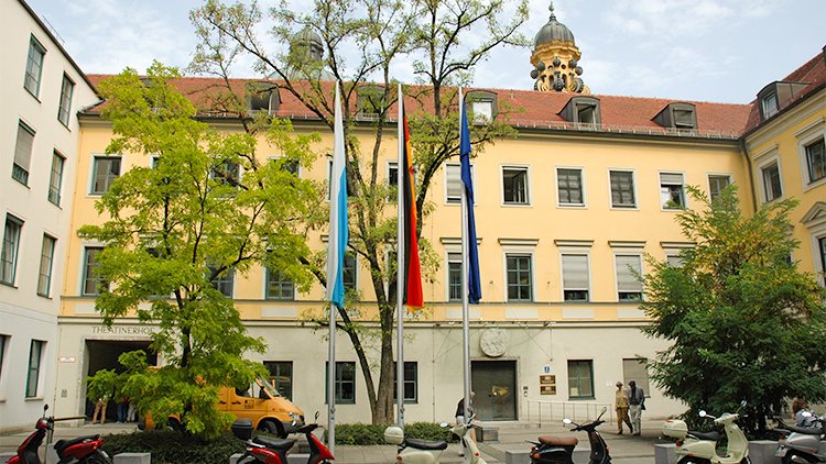 Gelbes Gebäude mit Deutschland-, EU- und Bayernflagge im Vordergrund.