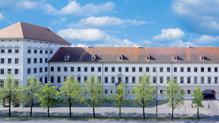 mehrstöckiges weißes Gebäude mit einer Baumreihe im Vordergrund.